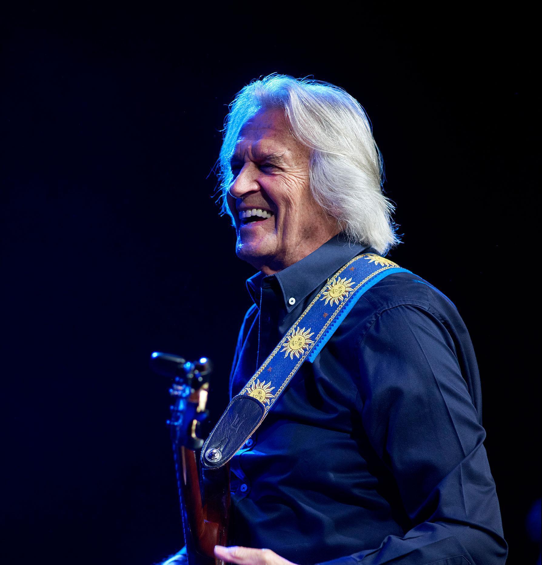 Profile shot of guitarist John McLaughlin with his instrument wrapped around him and laughing.