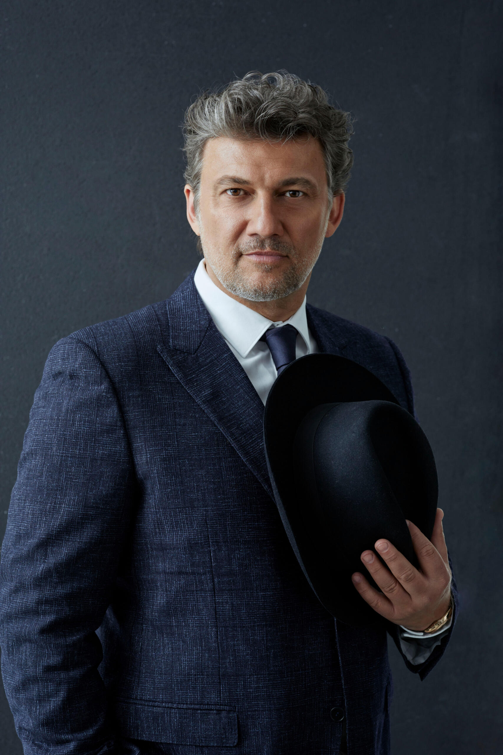 The singer Jonas Kaufmann stands in front of a dark wall. He is holding a hat in his hand.