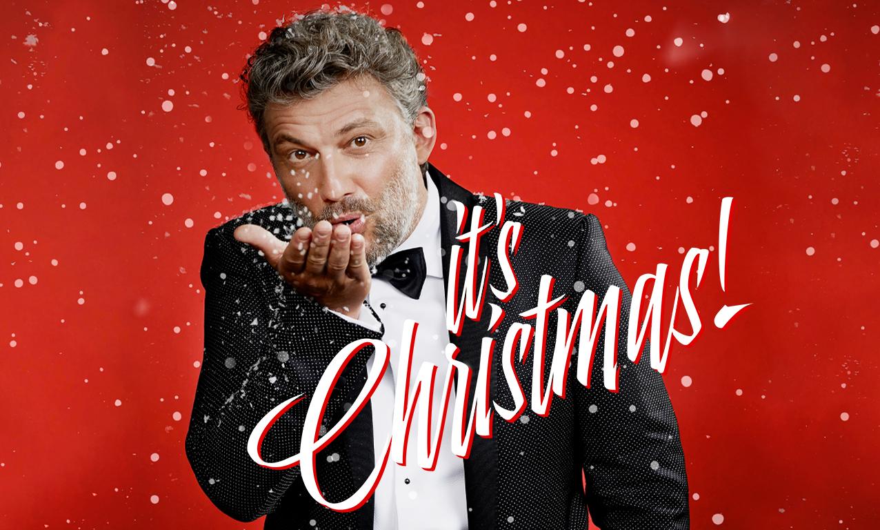 Tenor Jonas Kaufmann blows a few snowflakes in the direction of the person looking at him against a red background with white dots.