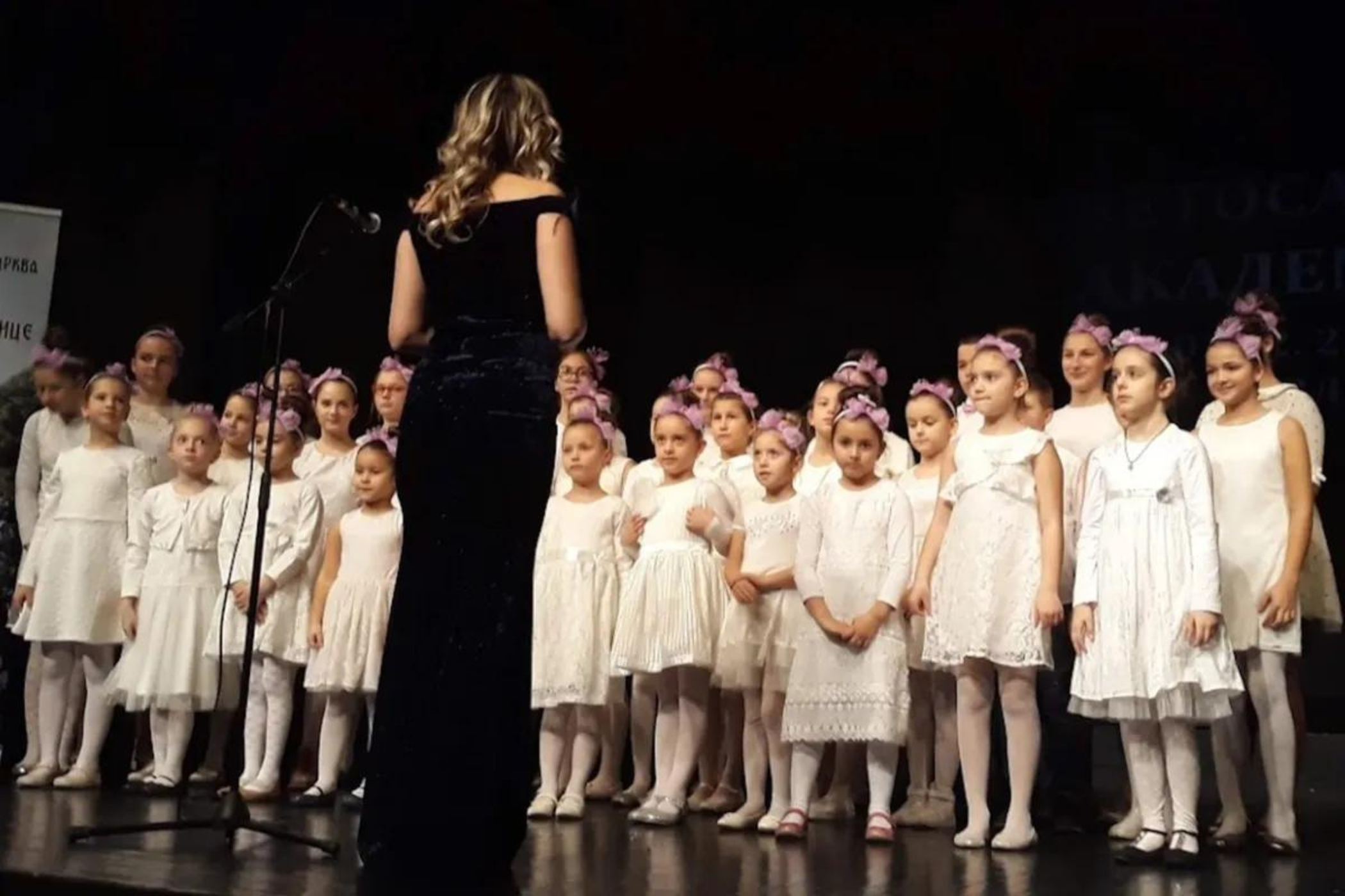 Many little girls in white dresses and pink bow on their heads are standing in a row on a stage, in front of them with their backs to us a woman in a long black evening dress.