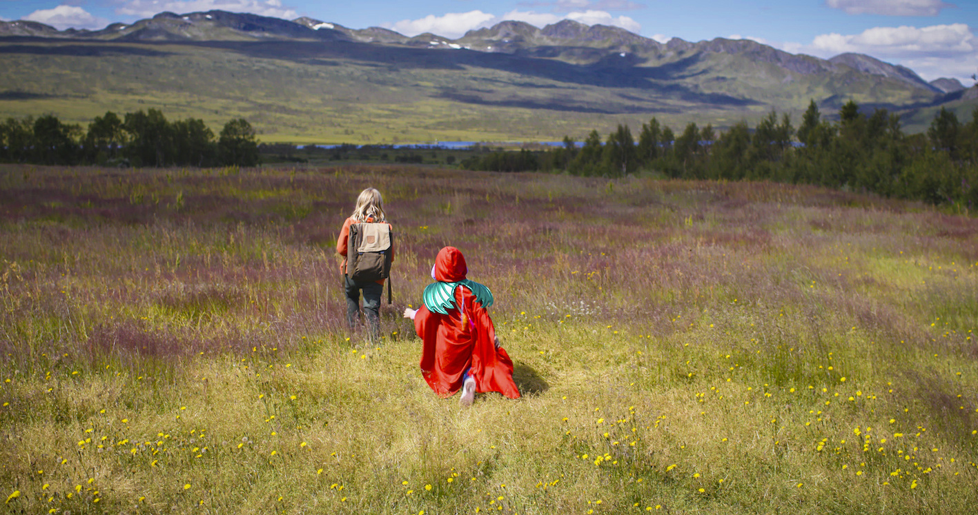 Zwei Kinder gehen über eine Blumenwiese, im Hintergrund sind Berge.