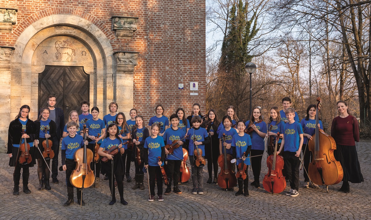 The square in front of a brick church. The musicians of the Children's Symphony stand on it with their string instruments.