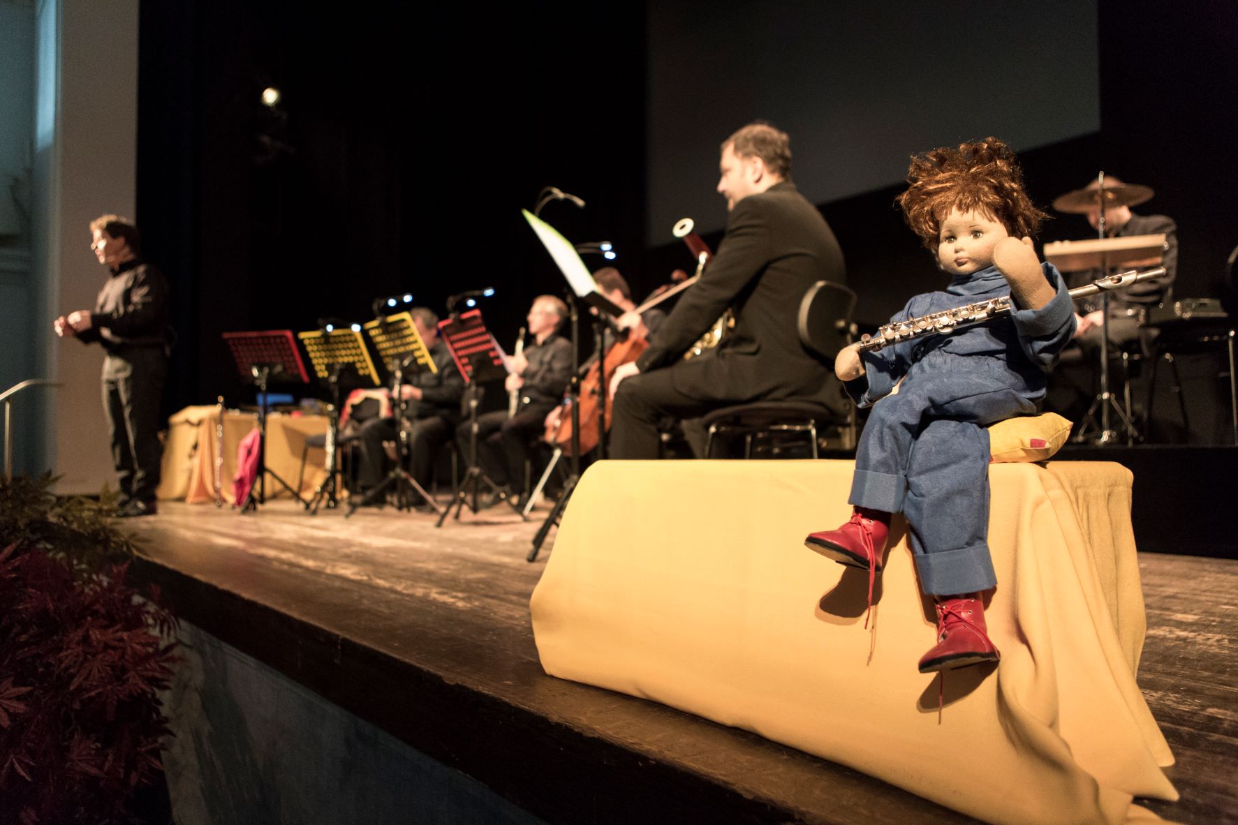 Eine Bühne mit Musikern von der Seite betrachtet, eine Marionette mit Querflöte sitzt am Rand