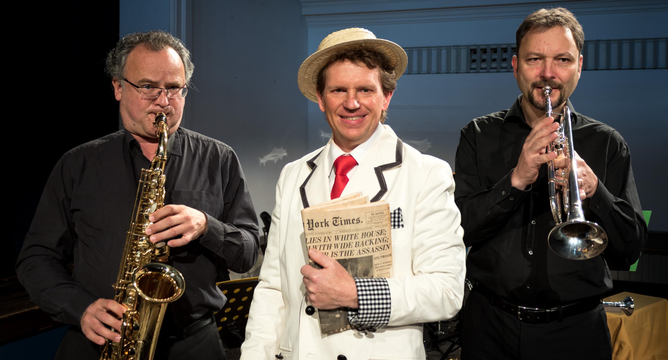 Drei Männer stehen nebeneinander. Rechts ein Trompeter, links ein Saxophonist und in der Mitte ein Mann in weißem Anzug und Hut mit einer Zeitung in der Hand.