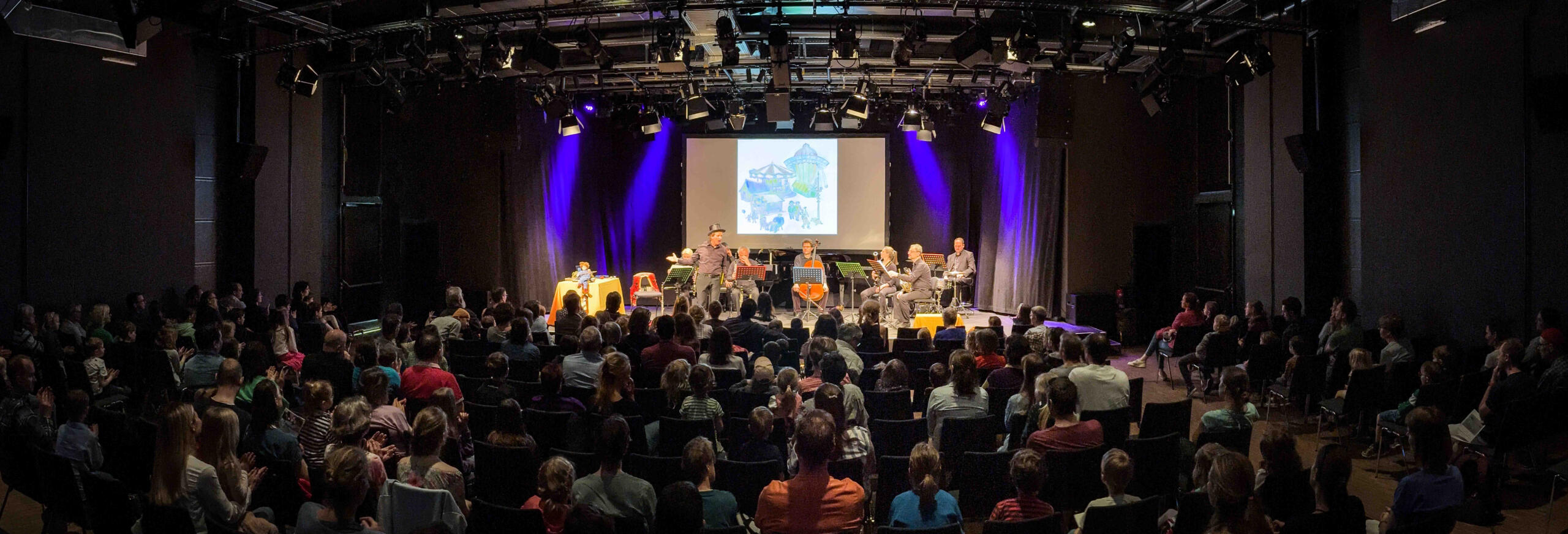 Panoramabild von einer Vorstellung aus dem Saal X am Gasteig HP8. Man sieht von hinten auf das Publikum und die hell erleuchtete Bühne auf der mehrere Schauspieler:innen und Musiker:innen sitzen.
