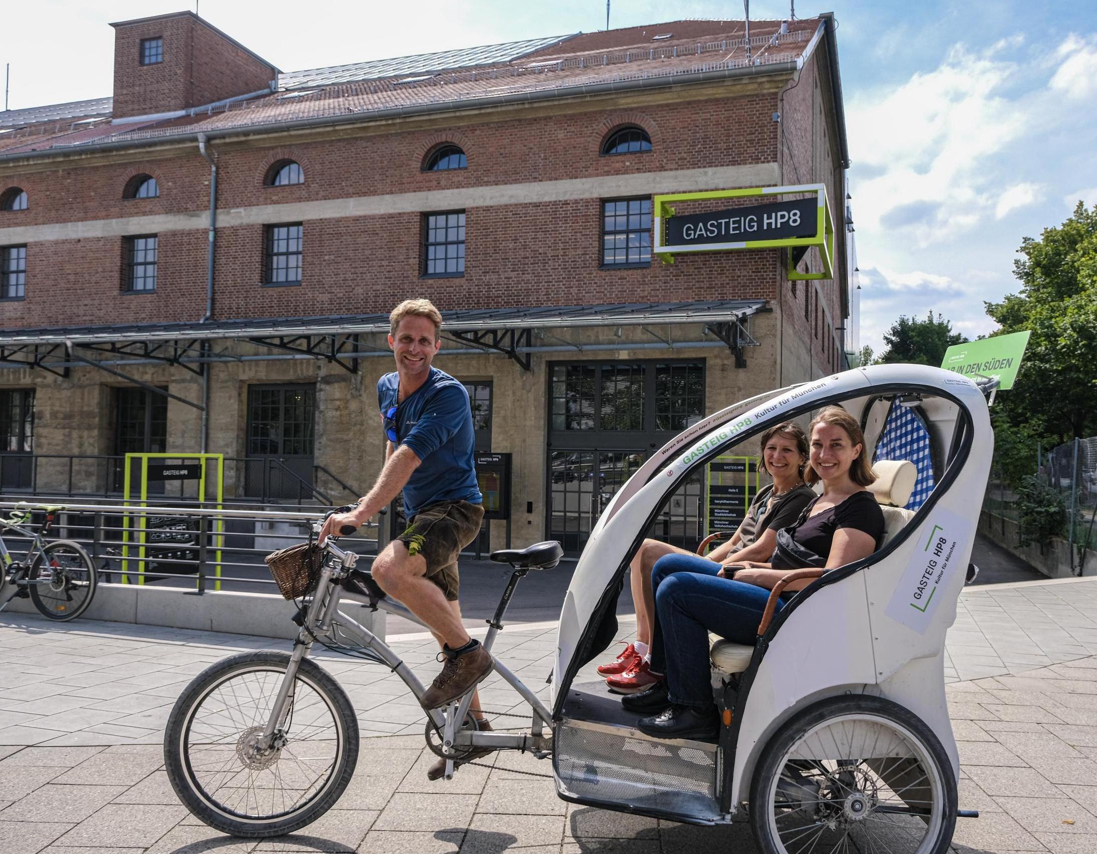 A bicycle rickshaw with female passengers in front of the brick facade of Hall E