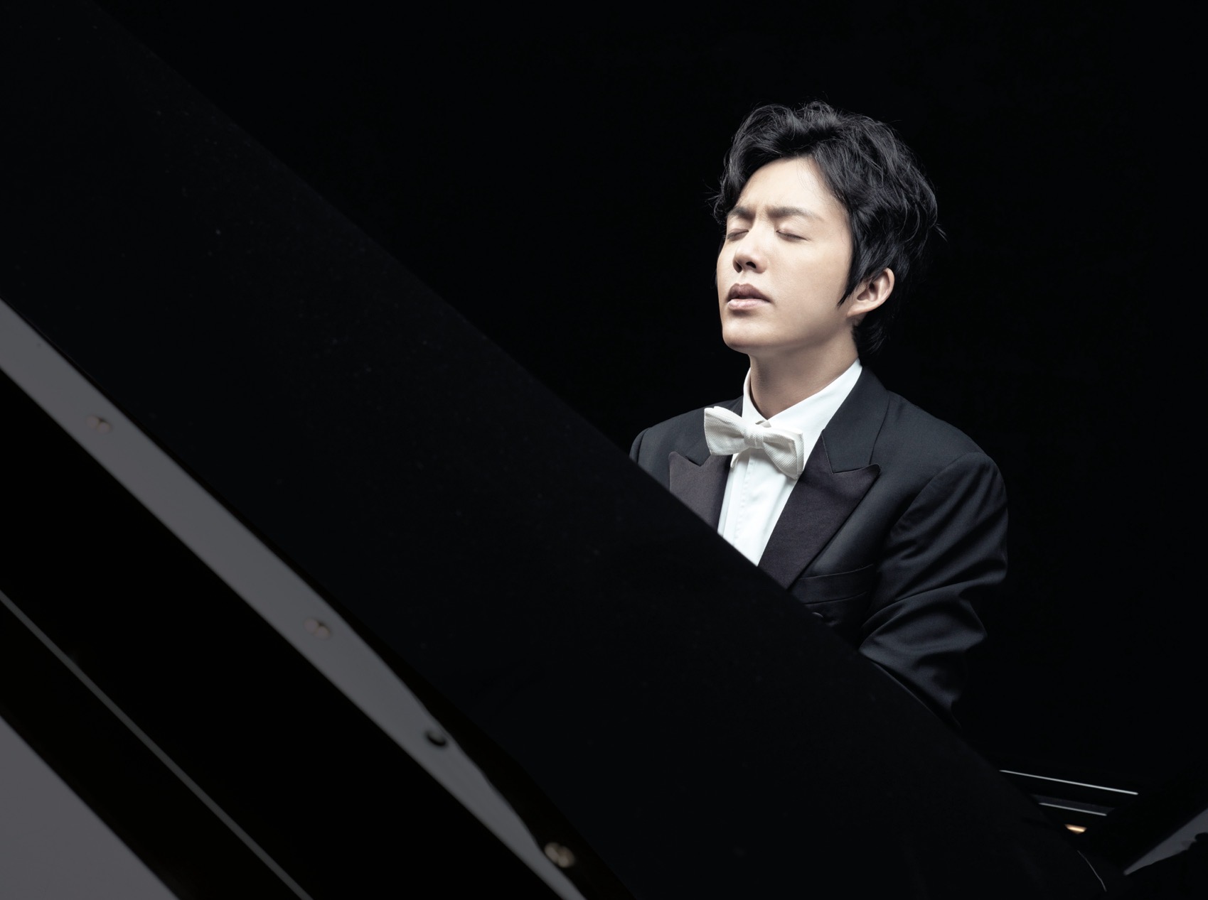A young pianist in a black dinner jacket plays the piano with his eyes closed.