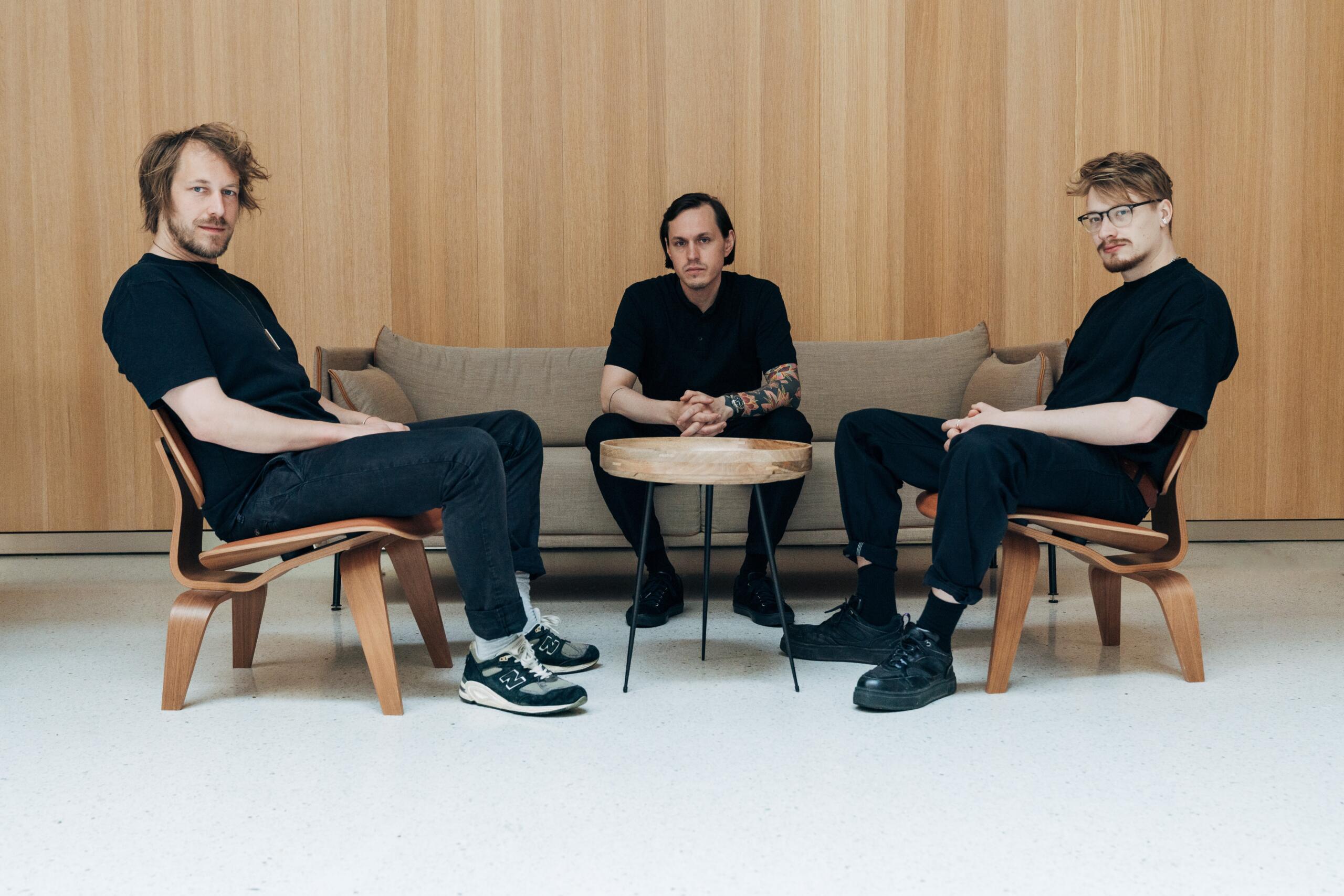 Drei Männer in schwarzer Kleidung sitzen um einen runden Holztisch und schauen in die Kamera. Der Raum ist hell und holzvertäfelt.