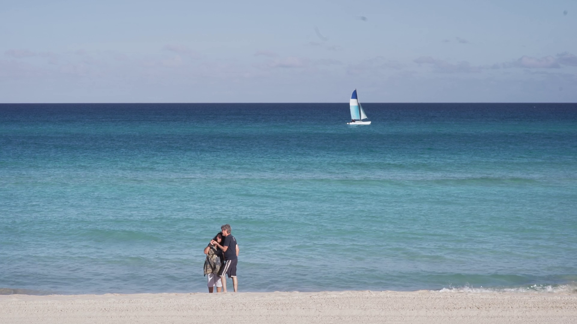 Zwei Personen stehen auf einem weißen Sandstrand direkt am Meer. Auf dem blauen Wasser fährt im Hintergrund ein Segelboot vorbei.