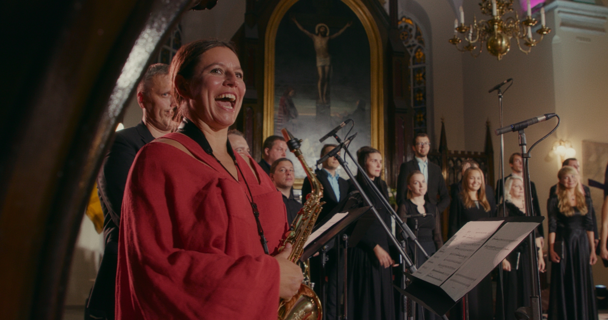 Still aus dem estländischen Film "Machina Faust". Man sieht einen Chor und eine Musikerin mit einem Saxophon in einer Kapelle stehen.