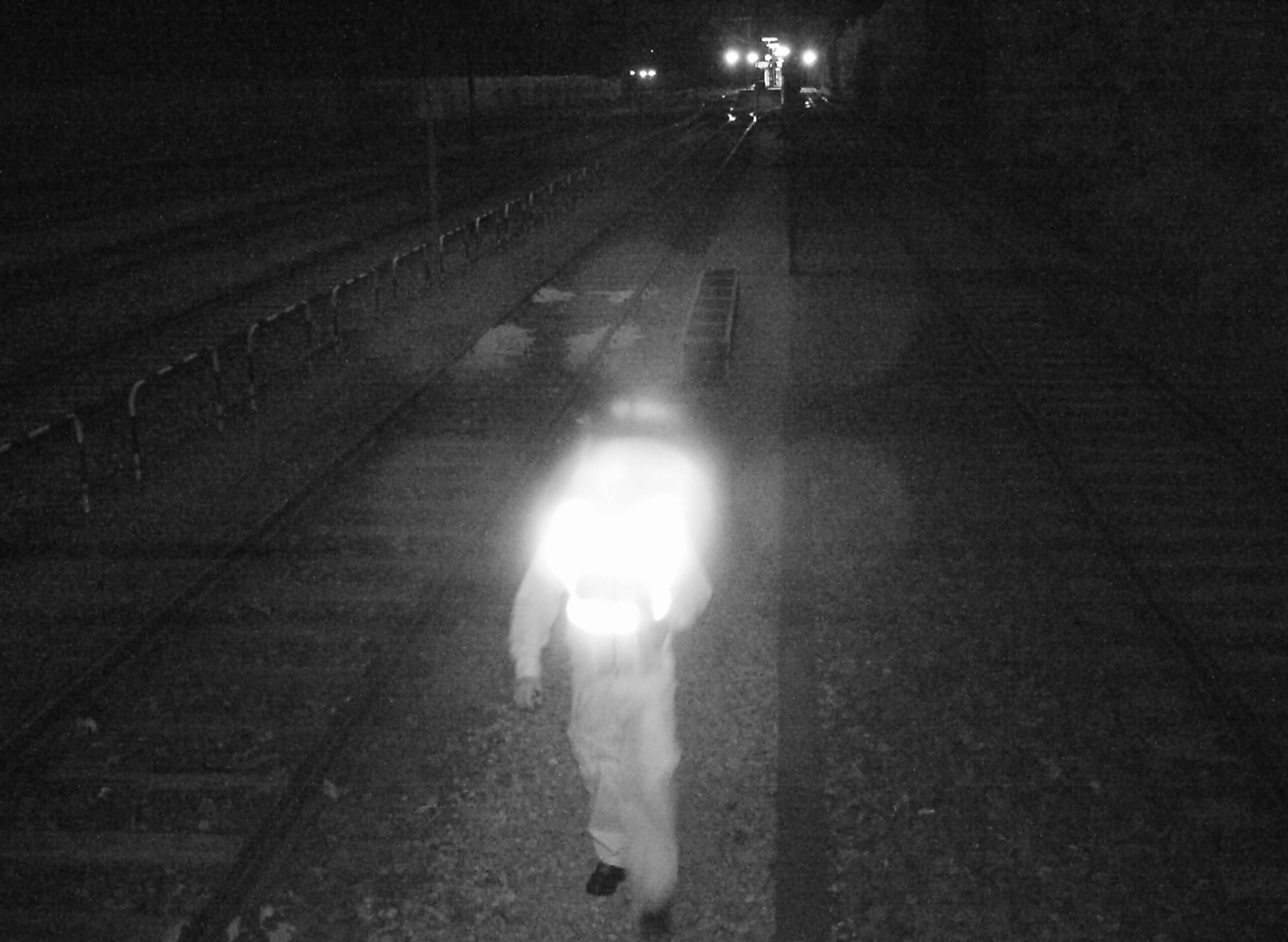 Nachtaufnahme, eine nicht zu erkennende Person in einem weißen Overall läuft an Bahngleisen entlang.