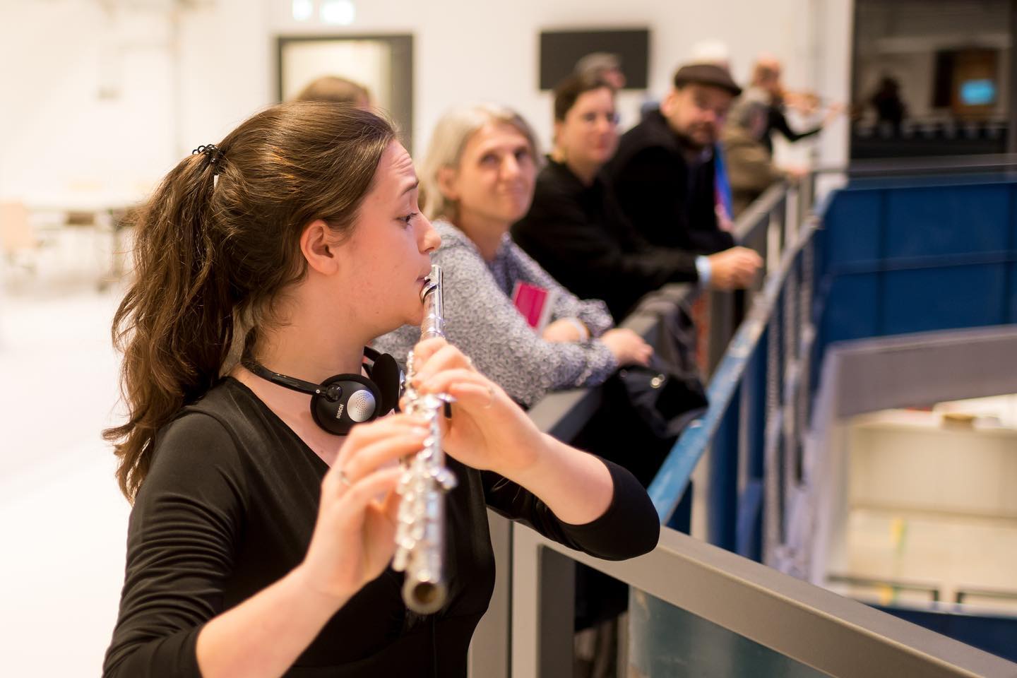 A young flutist at the ballustrade of Hall E, she plays, people watch next to her.
