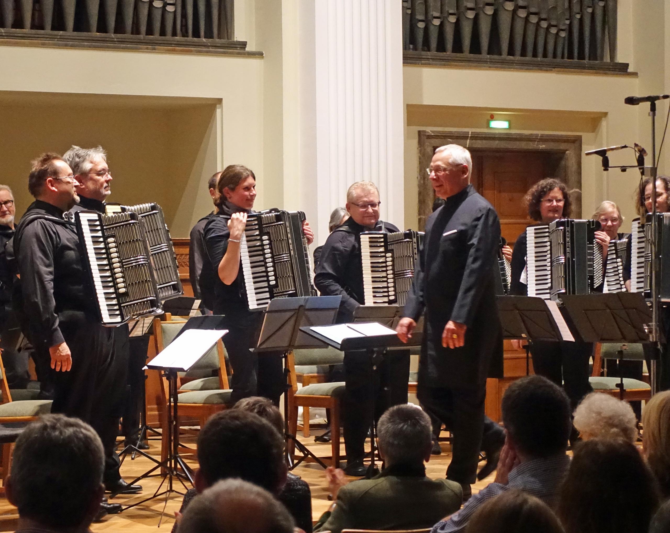 The accordion orchestra is seen during a concert applause. The musicians are standing behind their music stands, their accordions in their hands. In front of them you can see their conductor.