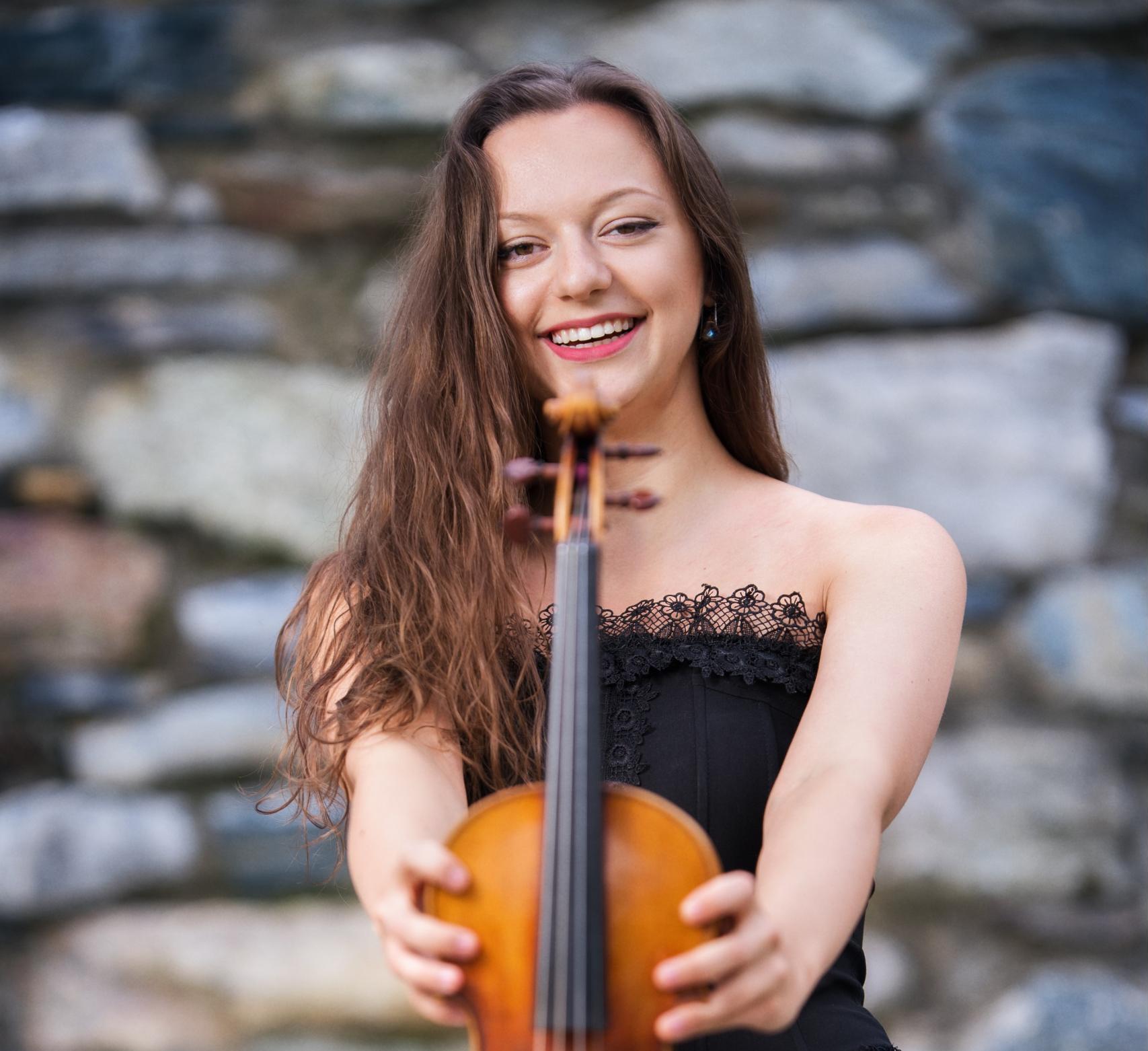 Violinist Maria Ioudenitch stands in front of a stone wall, laughing and holding her violin up to the camera.