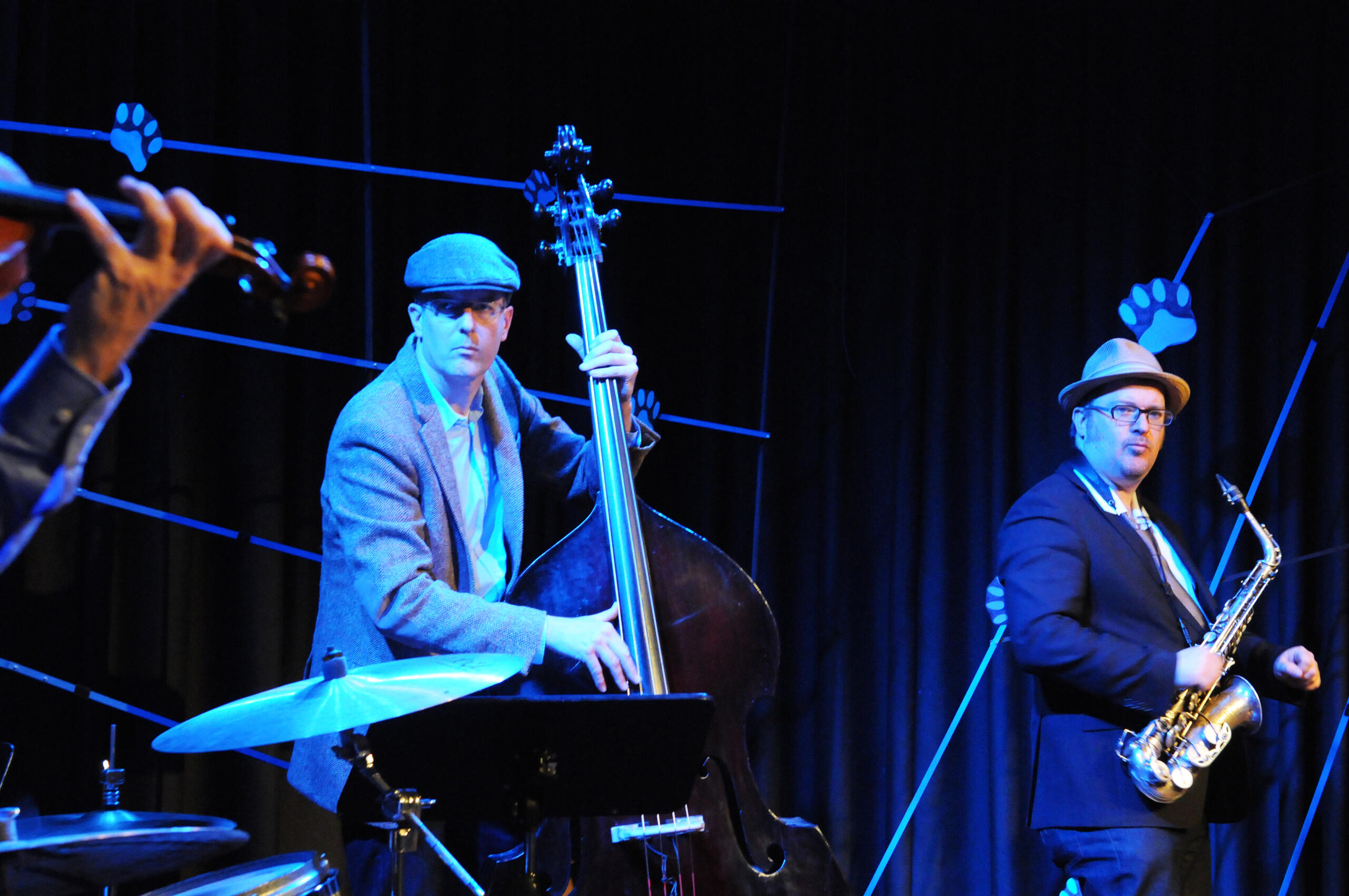 Ein Musiker mit einem Kontrabass und ein Musiker mit einem Saxophon stehen auf einer Bühne, die blau erleuchtet ist.