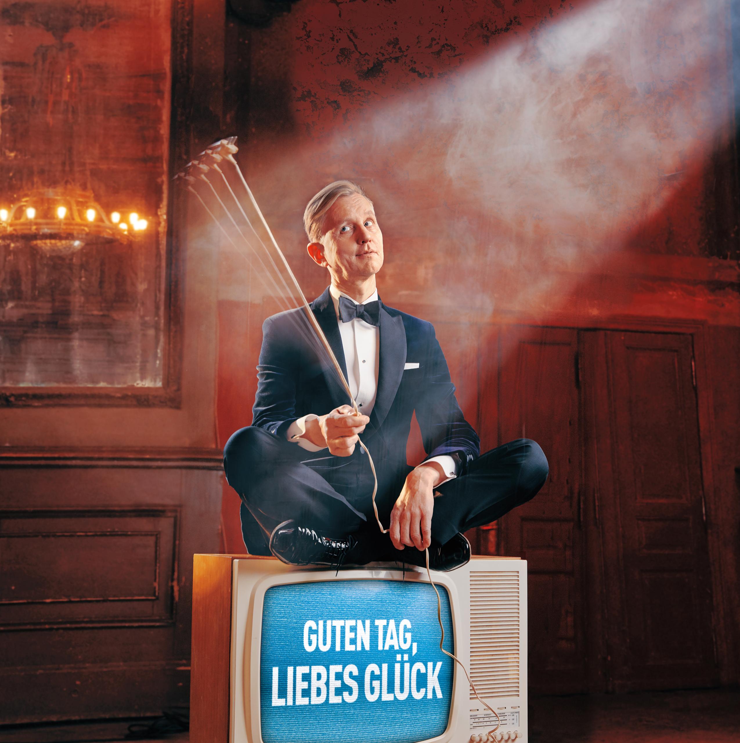 Max Raabe sits on an old TV, on the screen is Guten Tag liebbes Glück (Good day dear luck)
