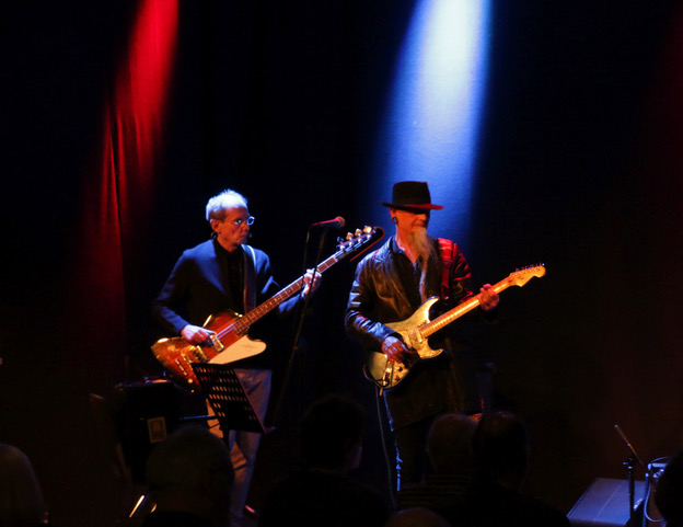 Eine Bühne mit blauem und rotem Sportscheinwerfer. Zwei Männer spielen E-Gitarre, einer trägt einen Hut.