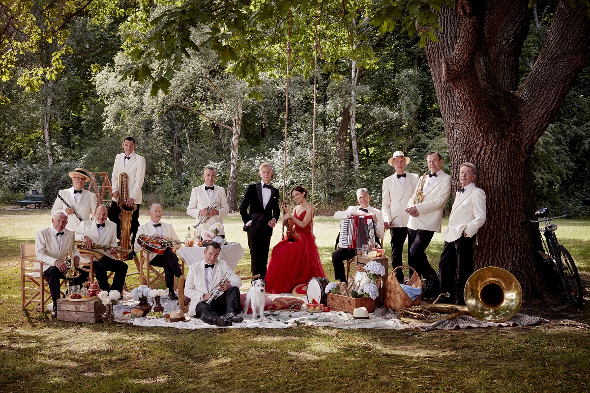 Max Raabe und das Palast Orchester stehen in Abendgarderobe auf einer Picknickdecke in einem sonnigen Park.