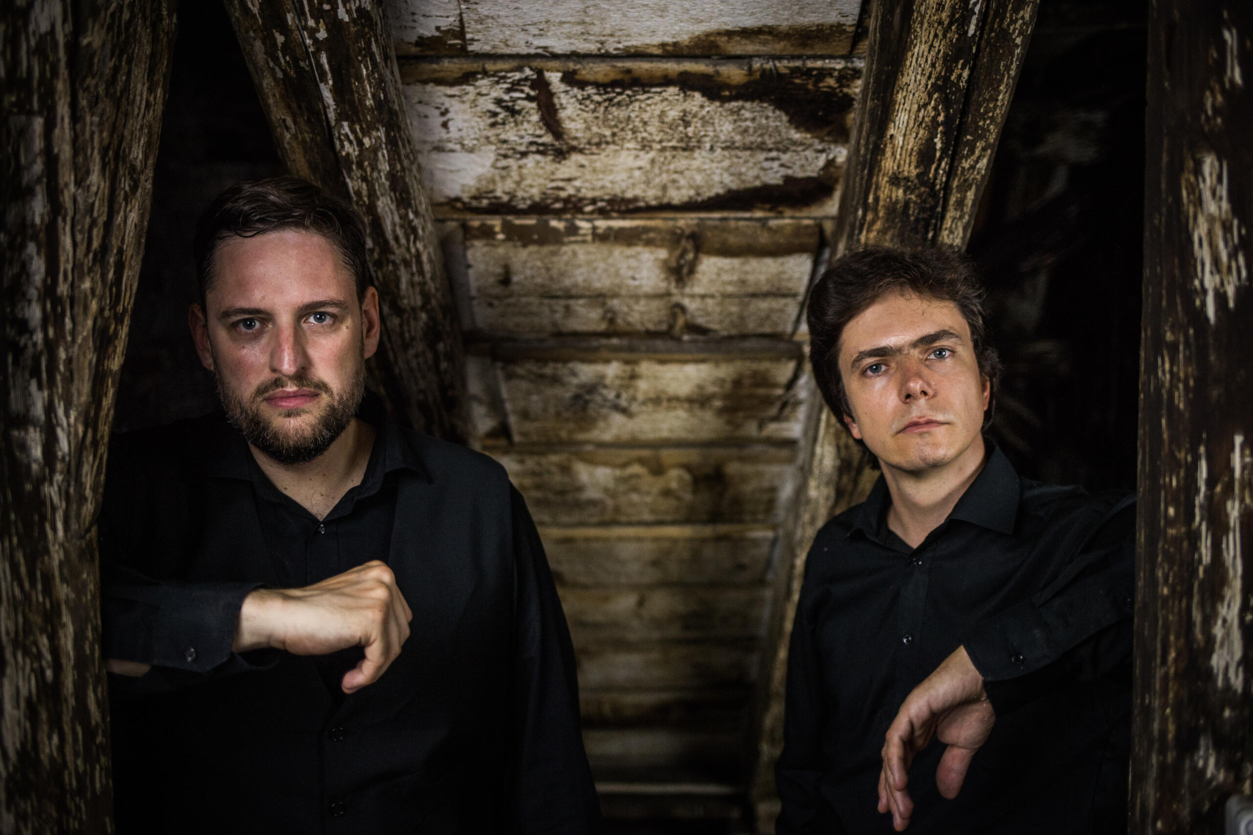 Two men dressed in black from a background of old wooden beams.