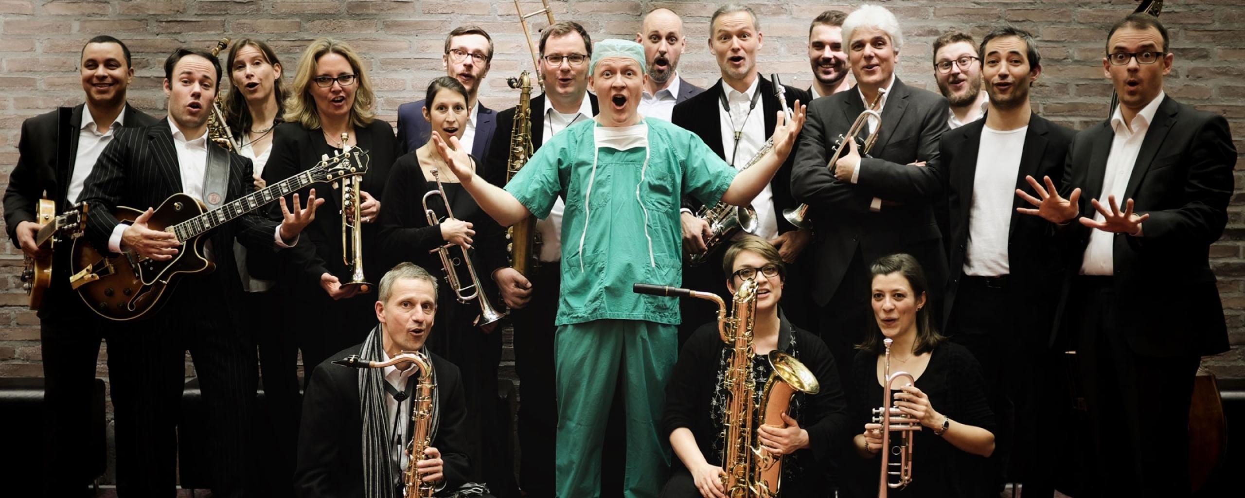 In der Mitte einer Gruppe von schwarz gekleideten Musiker*innen steht ein Arzt in grüner OP-Kleidung mit einem erstaunten Gesichtsausdruck.