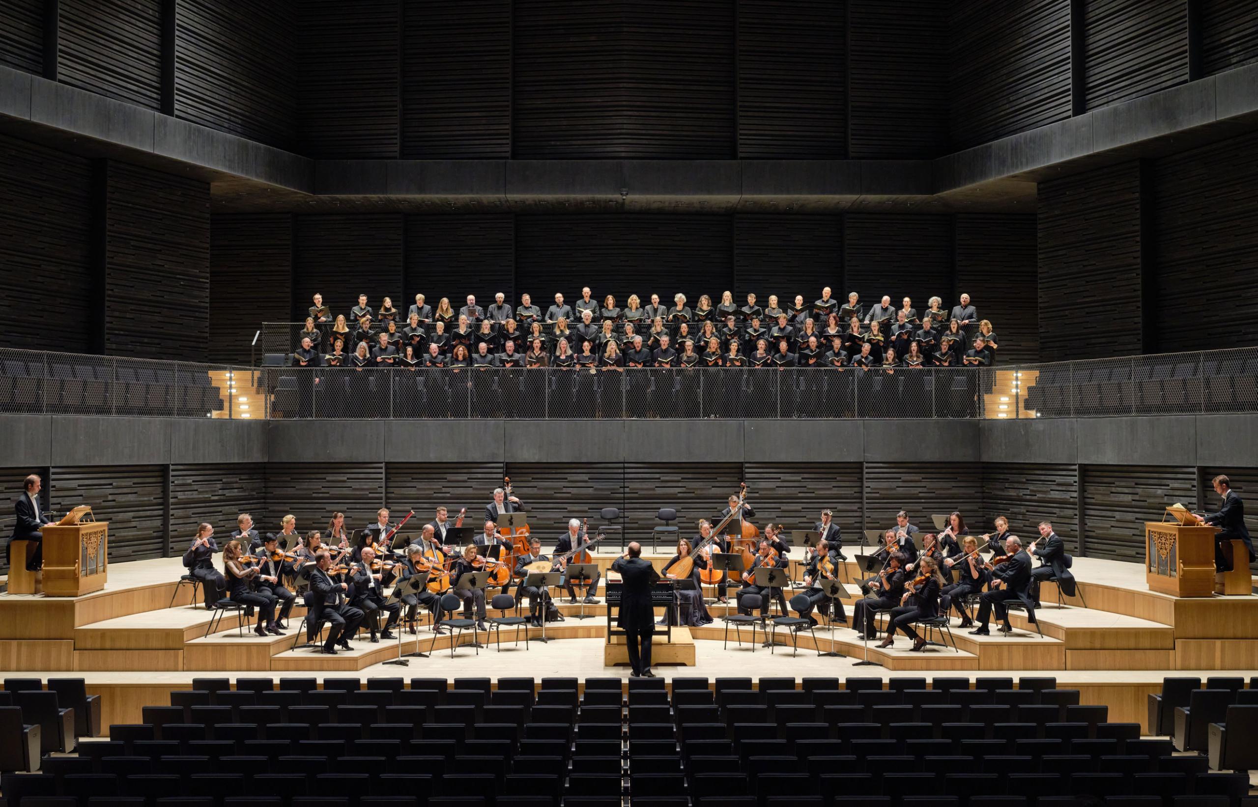 Der Münchener Bach-Chor ist in Position auf dem Chorbalkon in der Isarphilharmonie; auf der Bühne davor sieht man die Mitglieder des Münchener Bach-Orchesters mit Dirigent Hansjörg Albrecht.