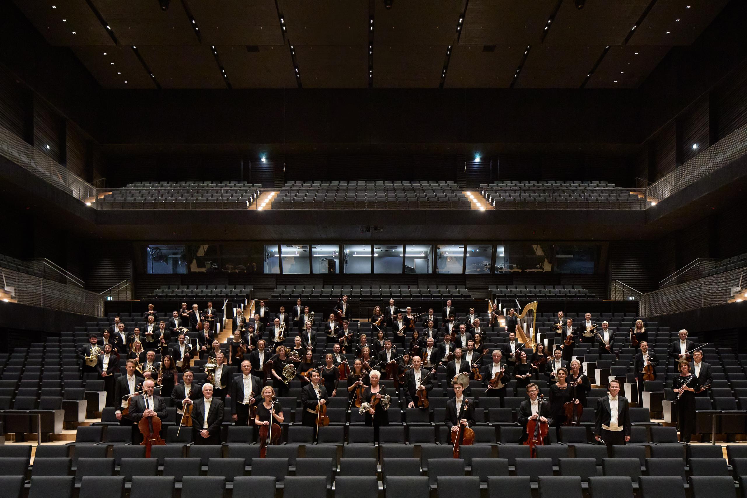 Dunkler Konzertsaal, Blick in den Zuschauerraum mit Sitzen und der Lichtregie. Die Musiker*innen stehen mit ihren Instrumenten zwischen den Sitzreihen