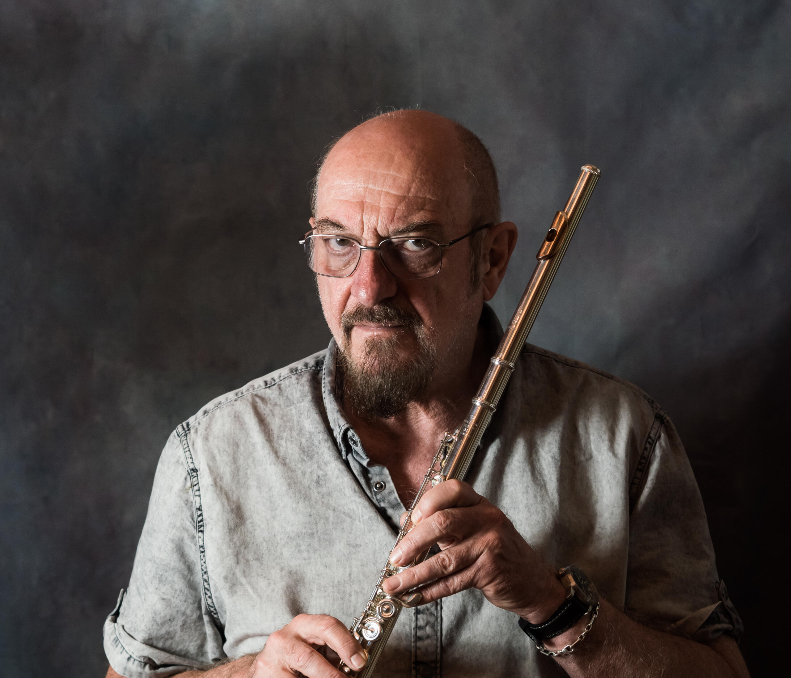Portrait photograph of the frontman of the band Jethro Tull, Ian Anderson. He is standing in front of a grey mottled background, holding his flute in both hands and looking seriously into the camera.
