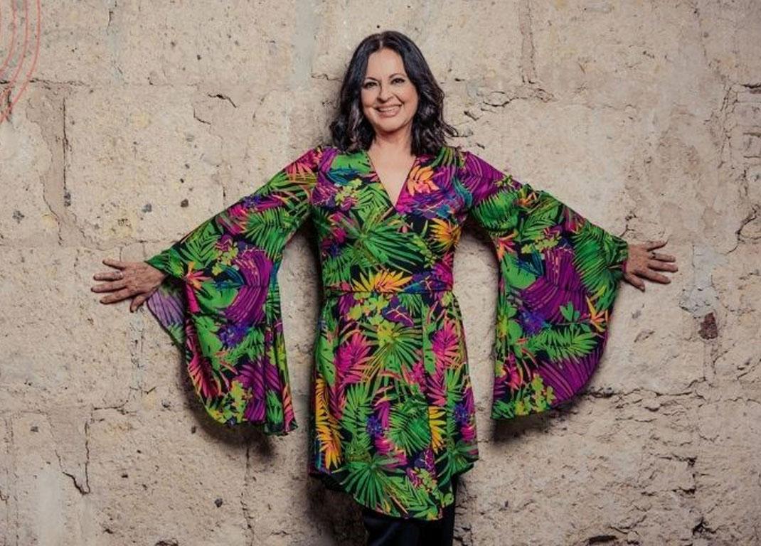 The singer Olga Cerpa is leaning against a sandstone wall with her arms spread out. She is wearing a colorful flower dress.