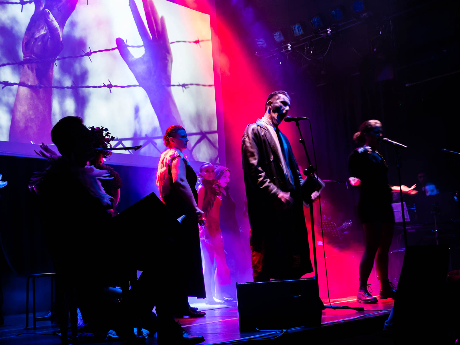 Eine Gruppe verkleideter Sänger und Sängerinnen steht auf einer Bühne, die in pinkes Licht getaucht ist. Im Hintergrund sieht man eine Projektion von Händen, die durch einen Stacheldrahtzaun greifen.