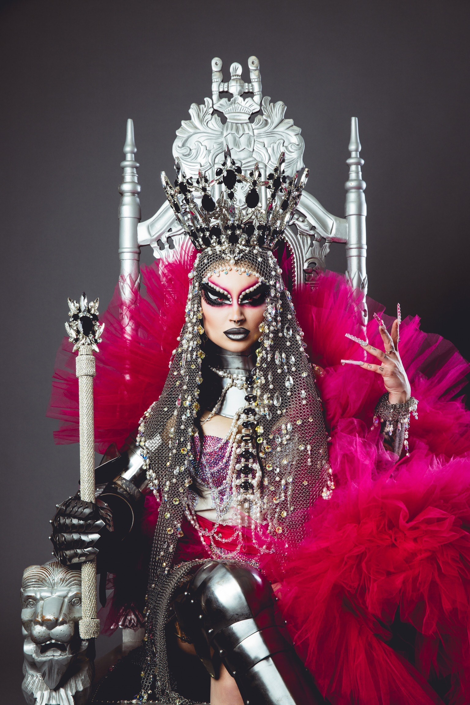 Eine Drag-Performerin sitzt auf einem Thron, trägt eine Krone und hält ein Zepter in der Hand.