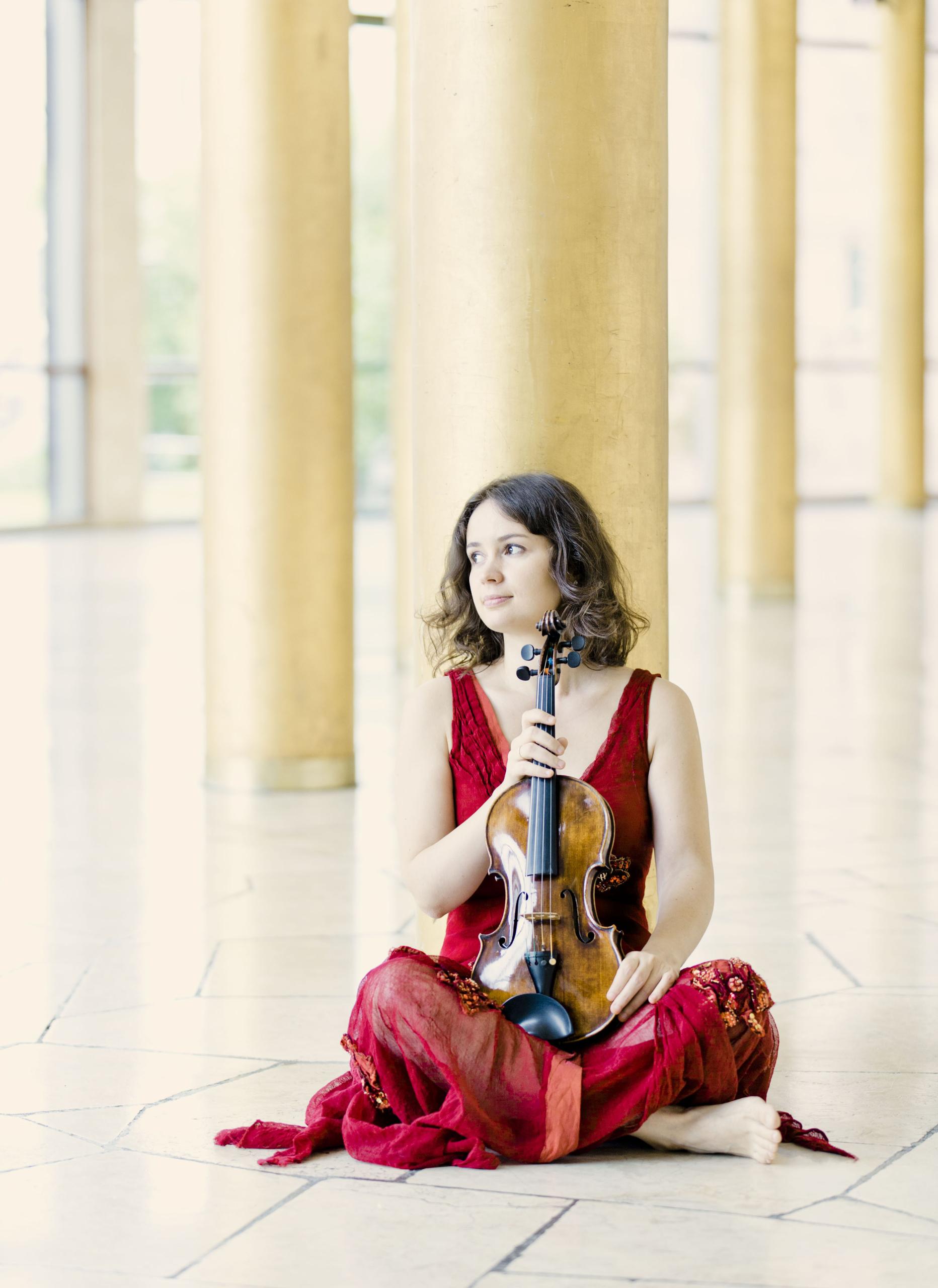 Violinist Patricia Kopachinskaya sits cross-legged on the floor and holds her violin on her lap