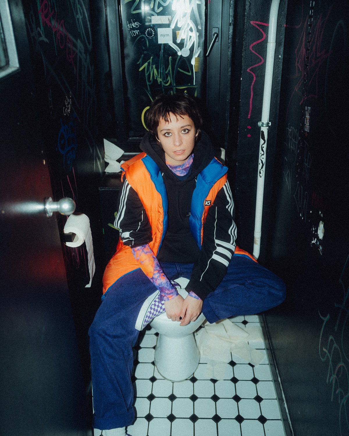 A young woman in athletic clothing sits on the toilet bowl of a pub. The walls are black and scrawled.