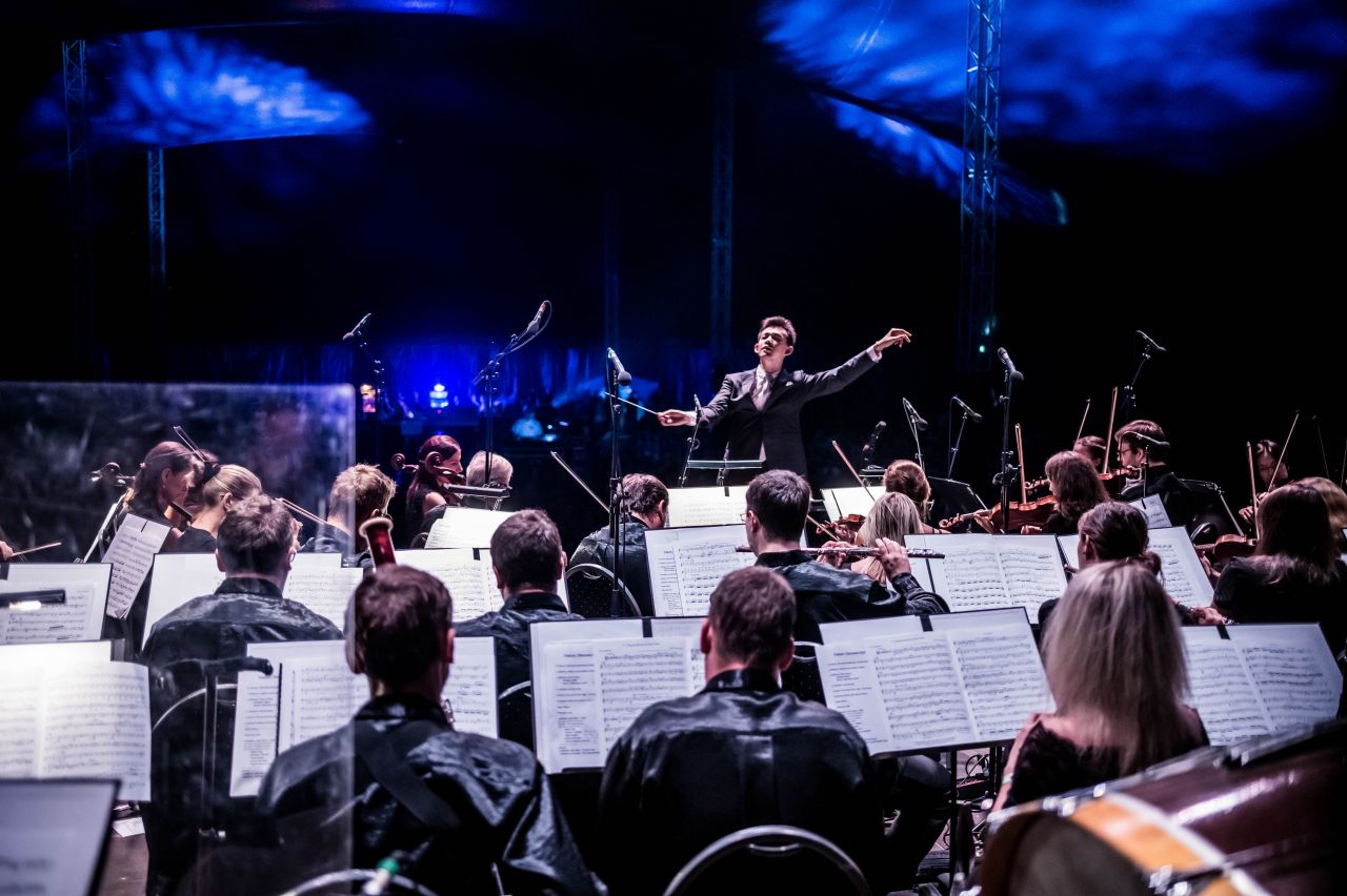 Musiker*innen beim Auftritt von hinten fotografiert, man sieht die Notenständer und einen Dirigenten in Aktion.