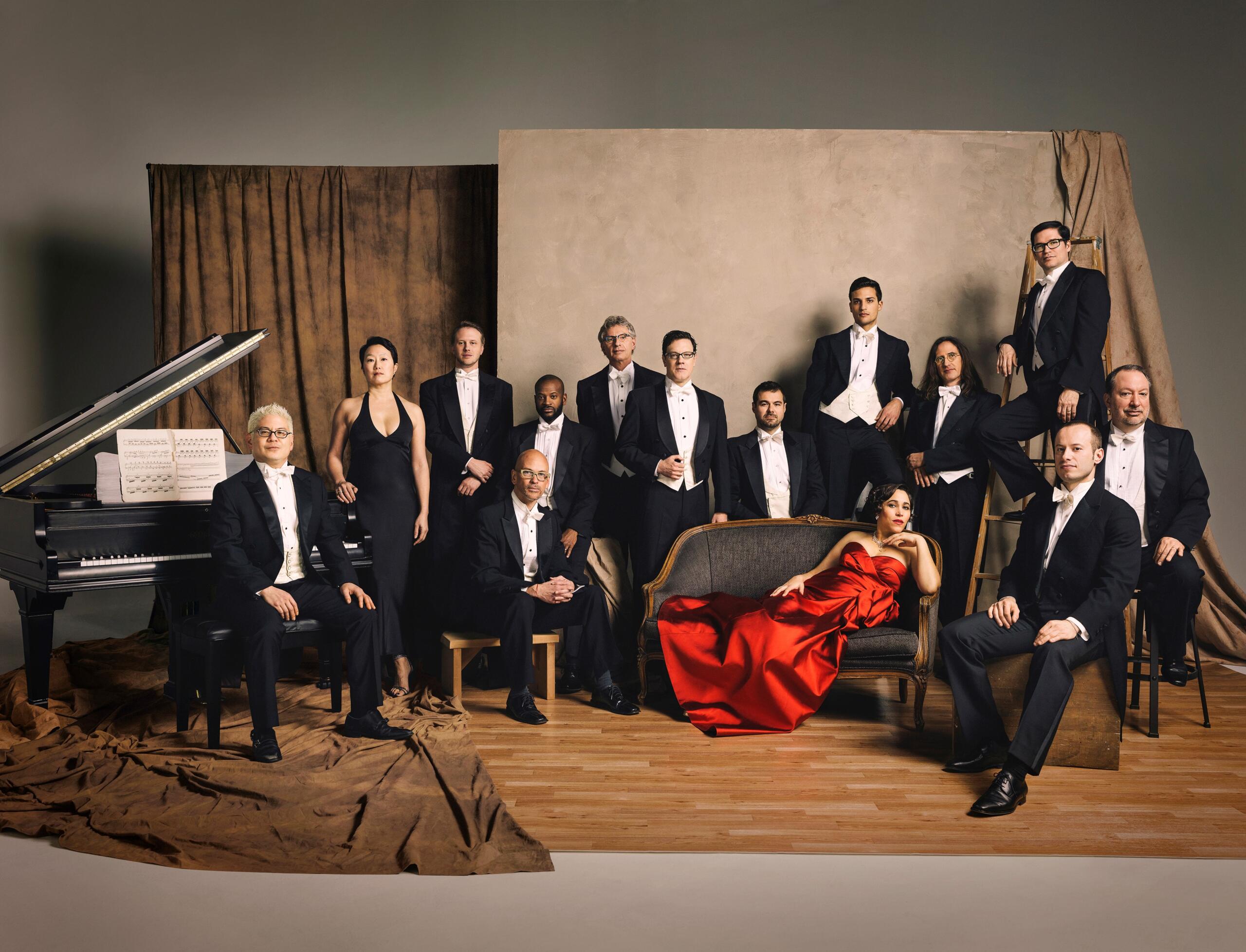 The Pink Martini orchestra sits, stands and lies against a beige background. Everyone wears black and white evening dress except for one singer in a red evening gown.