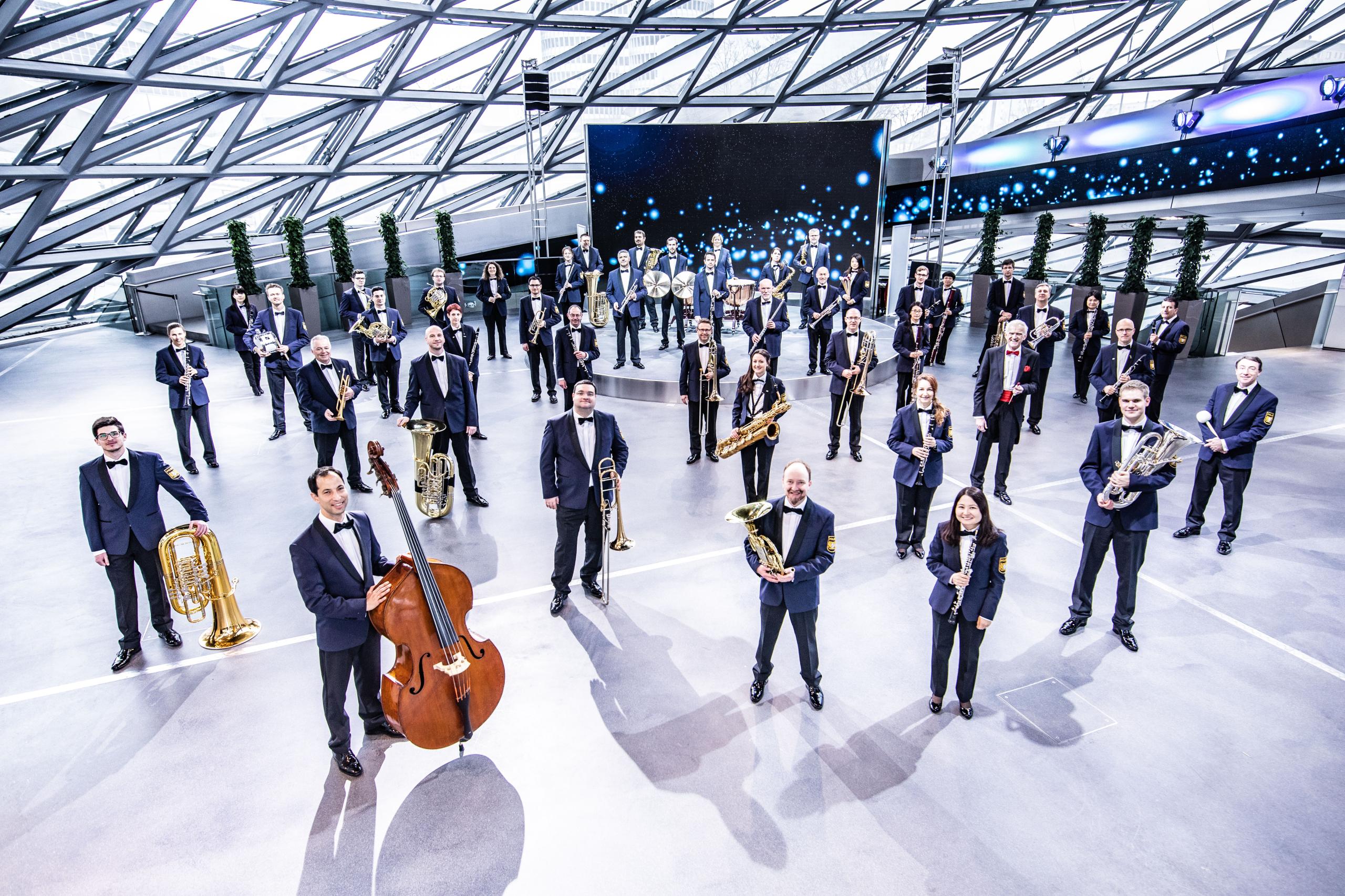 A glass hall in which musicians in blue suits are distributed individually. They each hold their instruments in their hands.