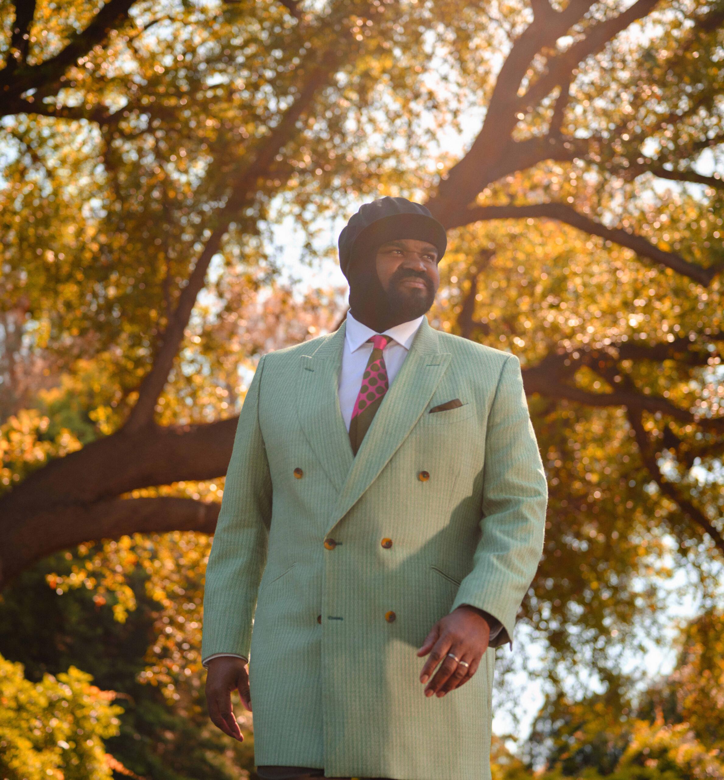 Portrait of the singer Gregory Porter. He is wearing a suit and standing in front of a large deciduous tree.