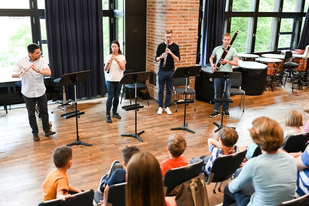 Ein Querflötist, eine Oboistin, ein Klarinettist und ein Fagottist spielen einer Gruppe Kinder mit Eltern etwas vor.