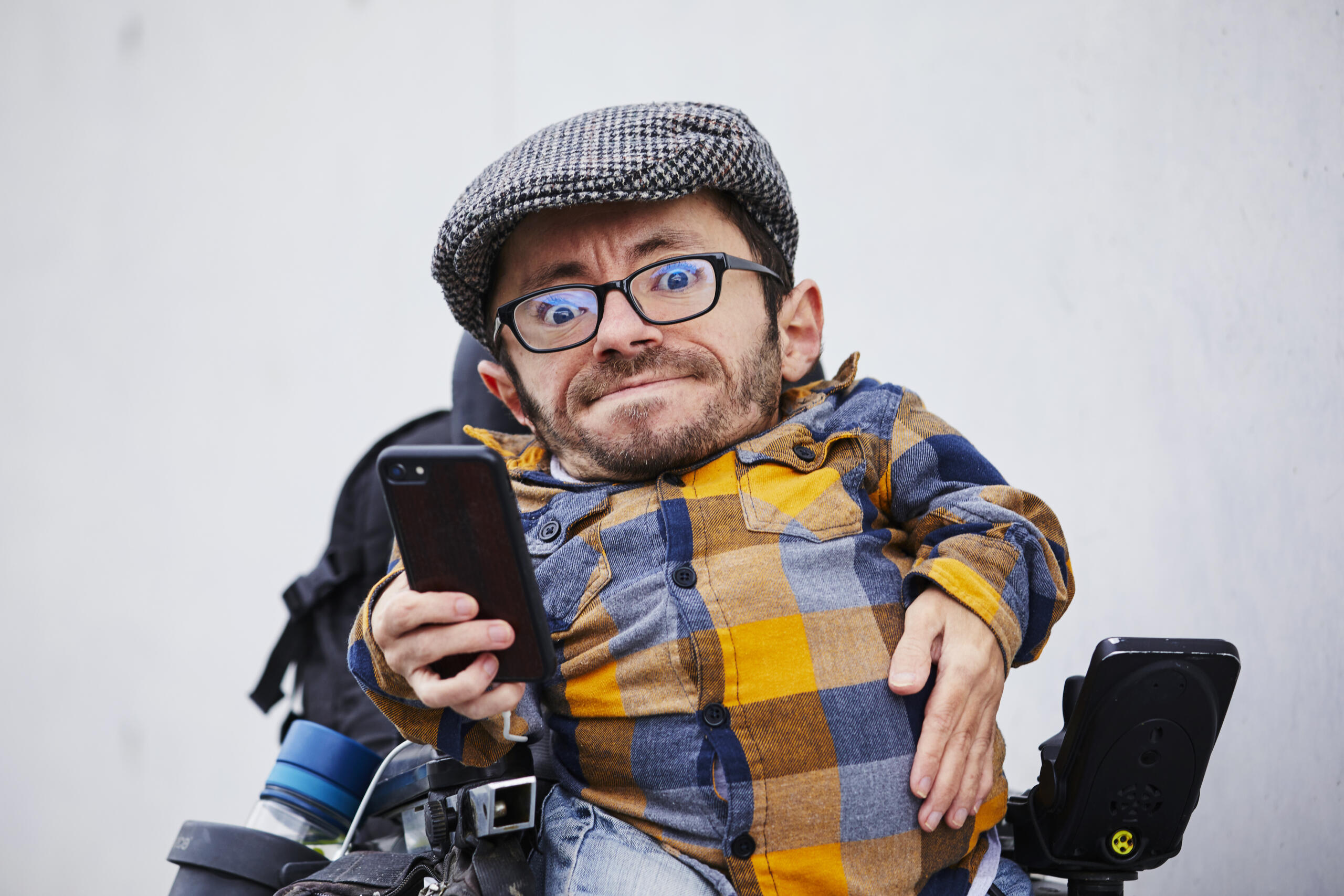 Ein Mann mit Brille, Bart und Schiebermütze. Er sitzt in einem Rollstuhl und bedient ein Smartphone.