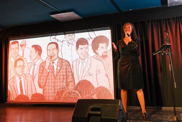 Eine Frau steht auf einer Bühne und spricht in ein Mikrofon. Neben ihr ist eine Leinwand auf der eine Illustration von einer Menschenmenge zu sehen ist.