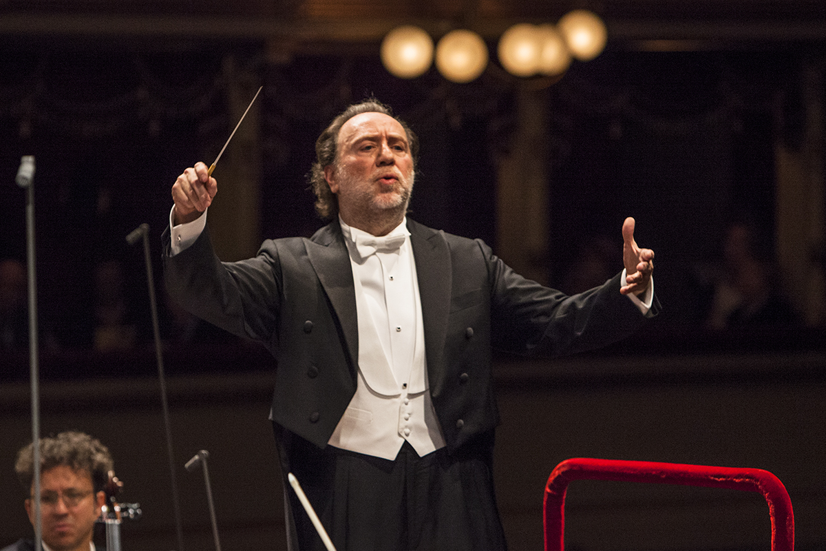 Der Dirigent Riccardo Chailly trägt einen schwarzen Frack mit einem weißen Hemd und dirigiert mit erhobenen Armen. In seiner rechten Hand hält er einen Taktstock.