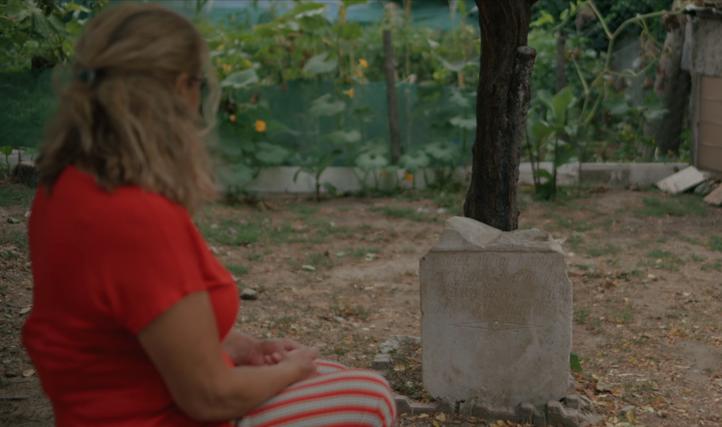 Eine Frau kniet vor einem schlichten Grabstein in einem Garten.
