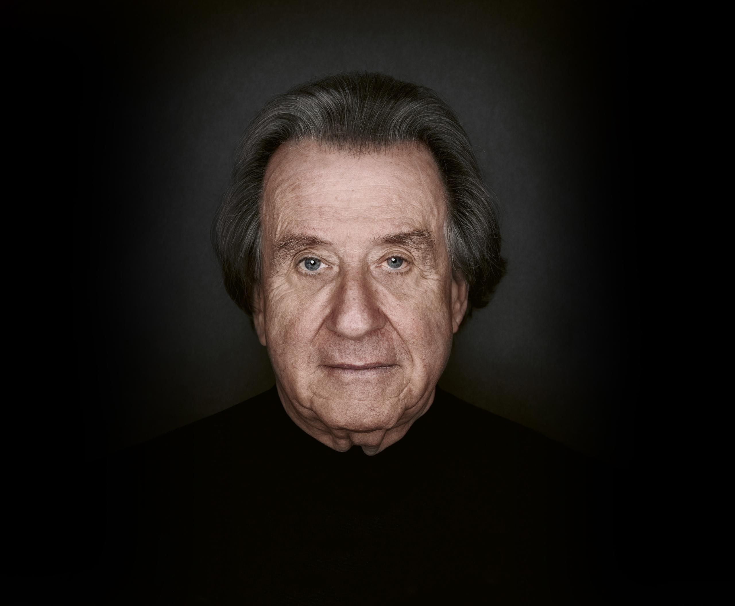 Portrait shot of the pianist Rudolf Buchbinder. In front of a dark background he looks directly into the camera.