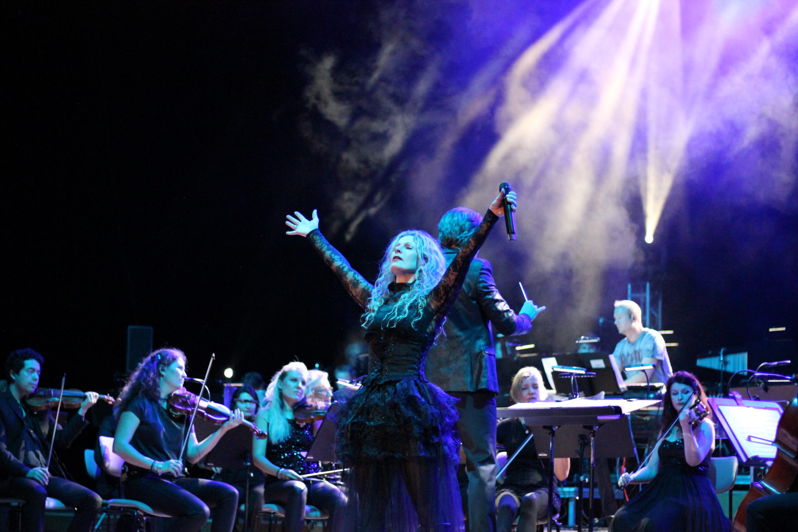 Eine Frau steht in einem schwarzen Kostüm auf einer Bühne, die blau erleuchtet ist. Im Hintergrund sitzen Musiker und Musikerinnen mit ihren Instrumenten.