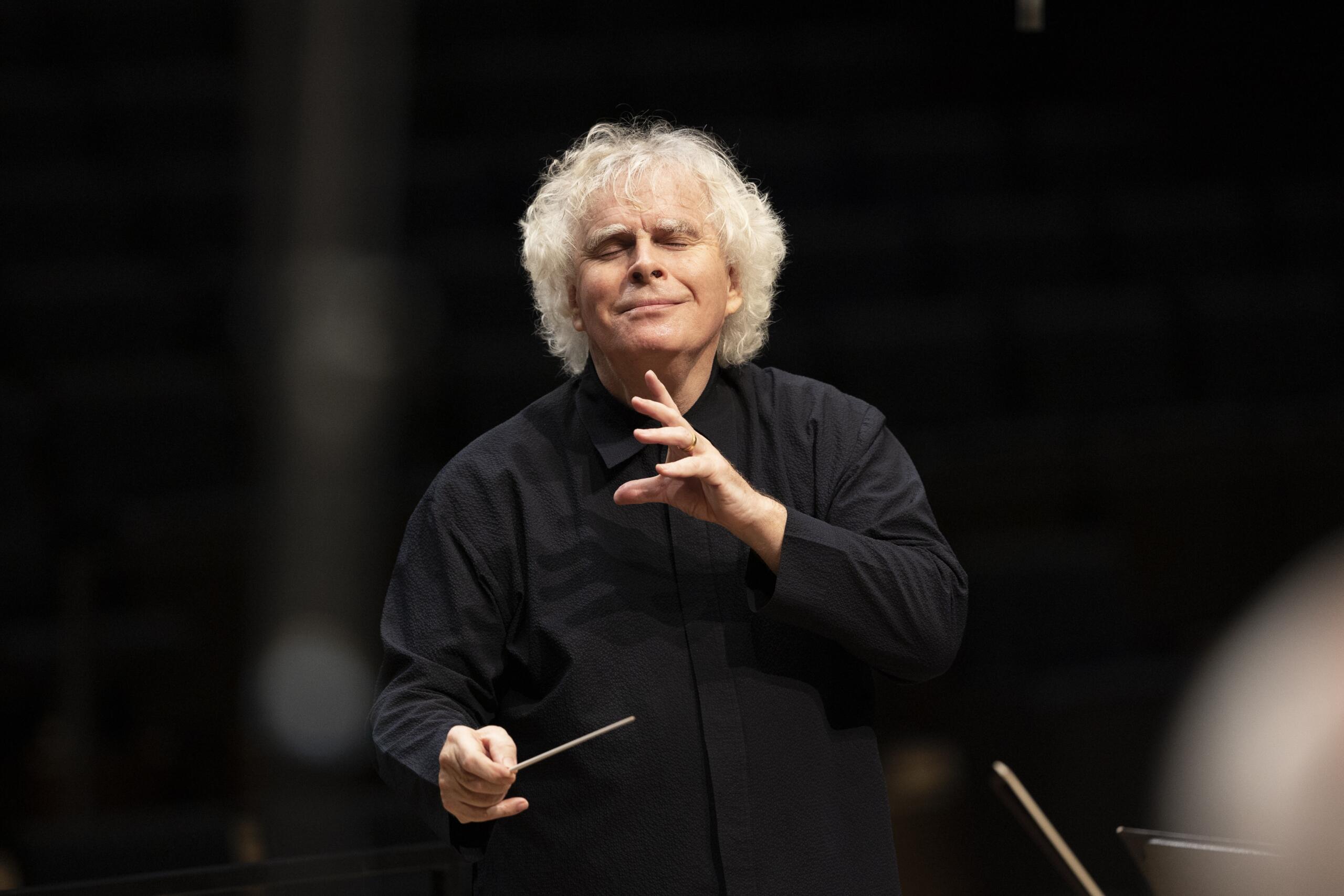 Conductor Rattle in black shirt conducting with his eyes closed.