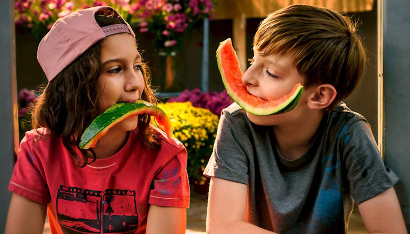 Zwei Kinder halten abgeknabberte Melonenstücken zwischen die Zähne geklemmt, so dass es aussieht, als würde das eine Kind lachen und das andere weinen.