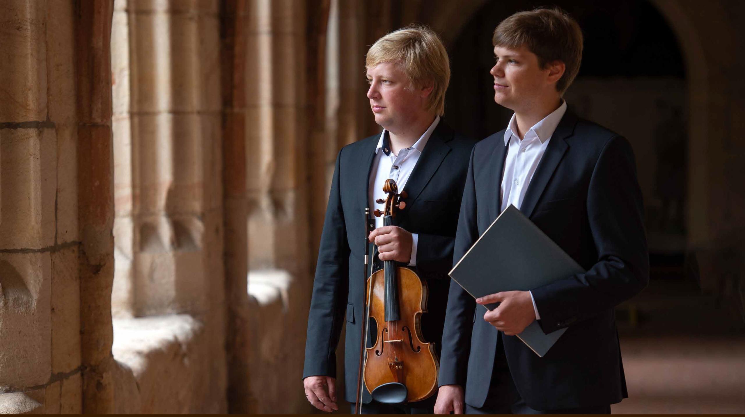 A historical cloister, two young musicians in dark suits, one with a violin, one with a music book
