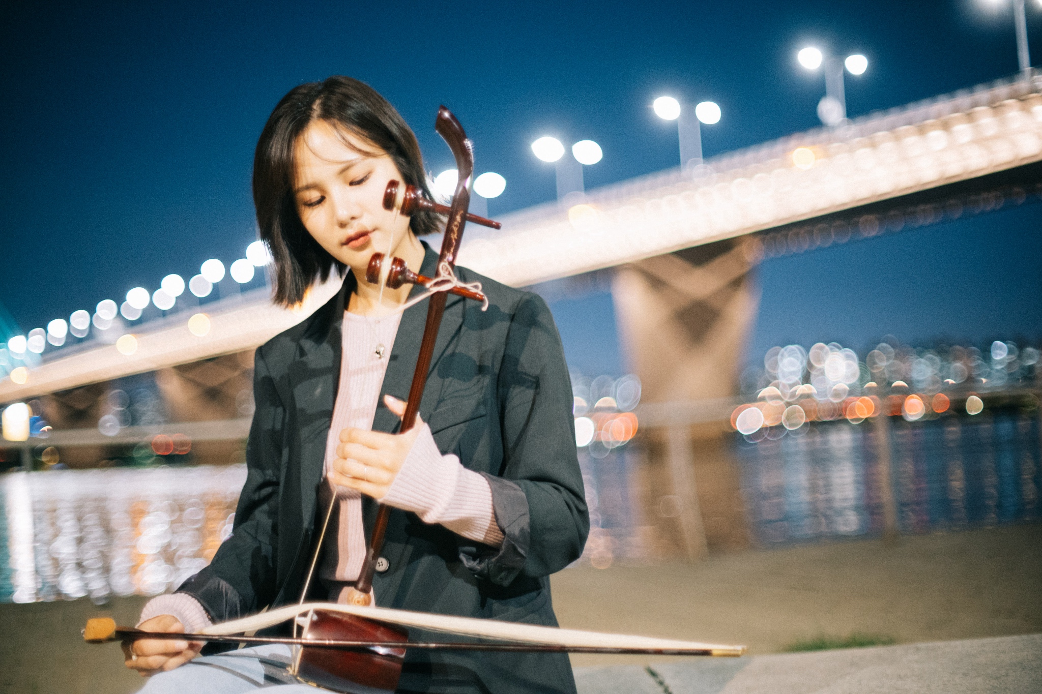 A young woman plays a stringed instrument. A car bridge and illuminated city in the background.