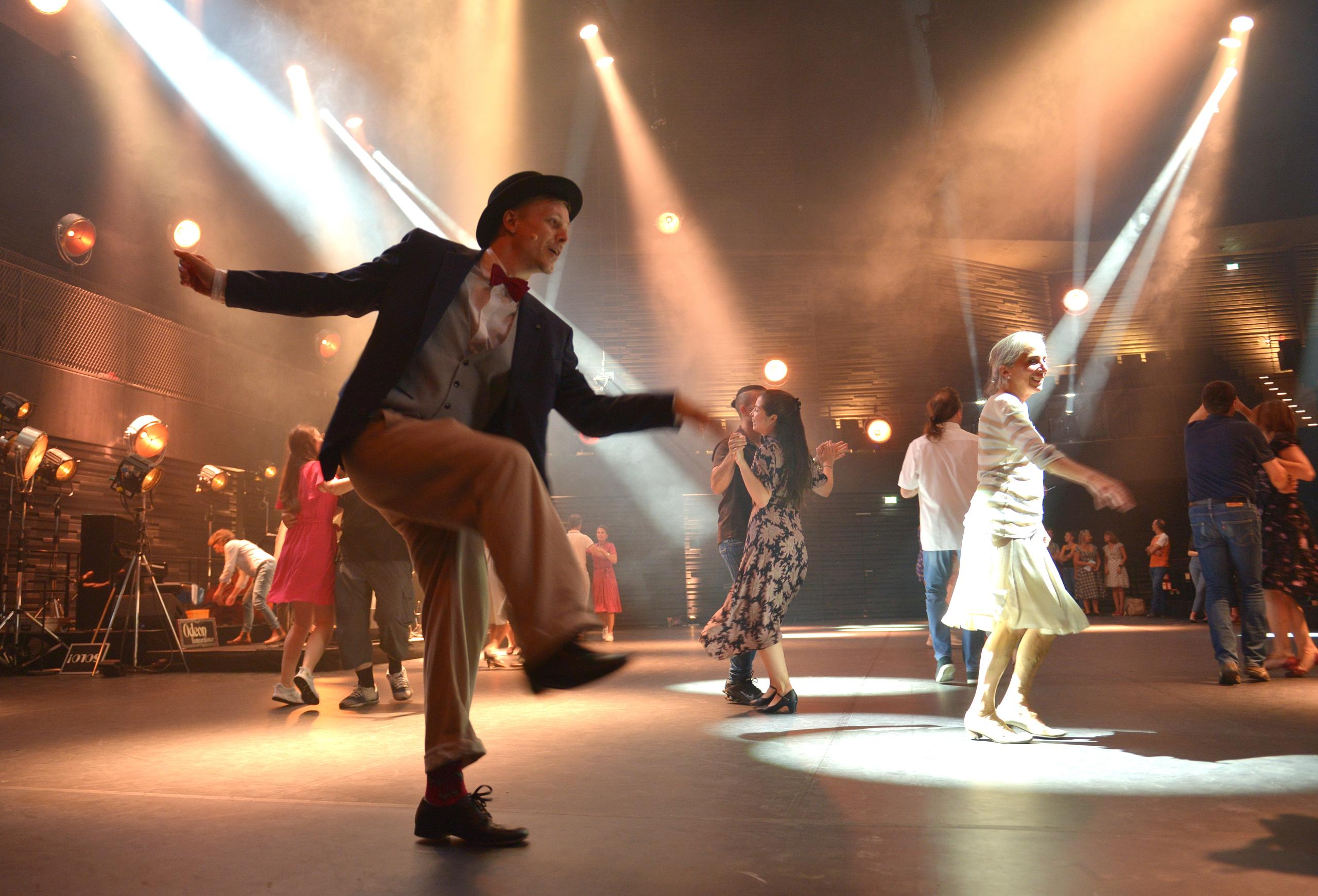 Tanzende Menschen auf der Bühne der Isarphilharmonie. Ein Mann mit Hut tanzt Swing im Vordergrund