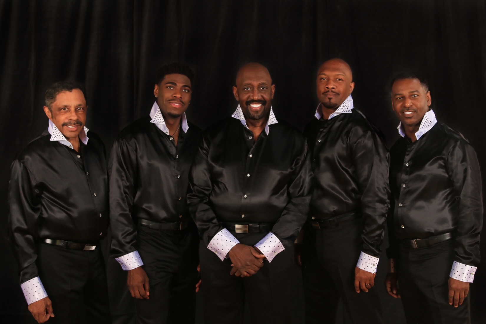 Die 5 Musiker der Temptations vor schwarzem Hintergrund, sie tragen schwarze Satinhemden mit weißen Krägen und Manschetten