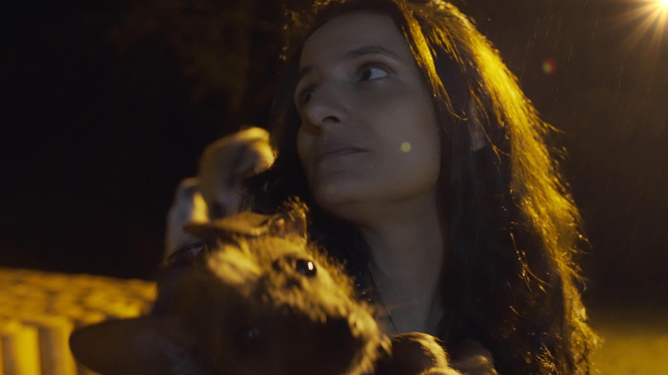 Ein Frau mit langen dunklen Haaren hält einen kleinen Hund auf dem Arm. Sie schaut zur Seite und wird von hinten von einer hellen Laterne angeleuchtet.